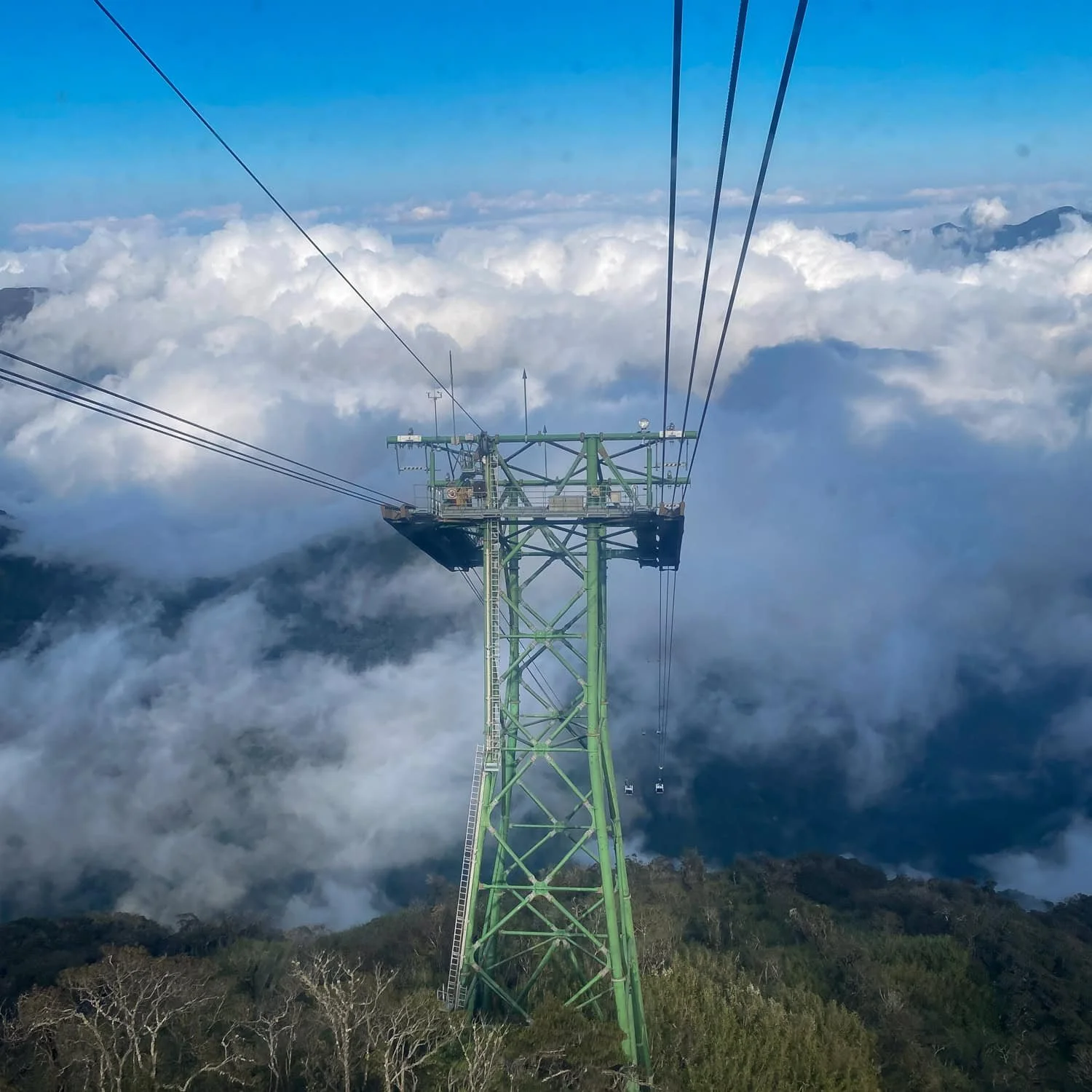 A tall green tower supports the Fansipan cable car lines above a dramatic sea of clouds and mountainous forest below.