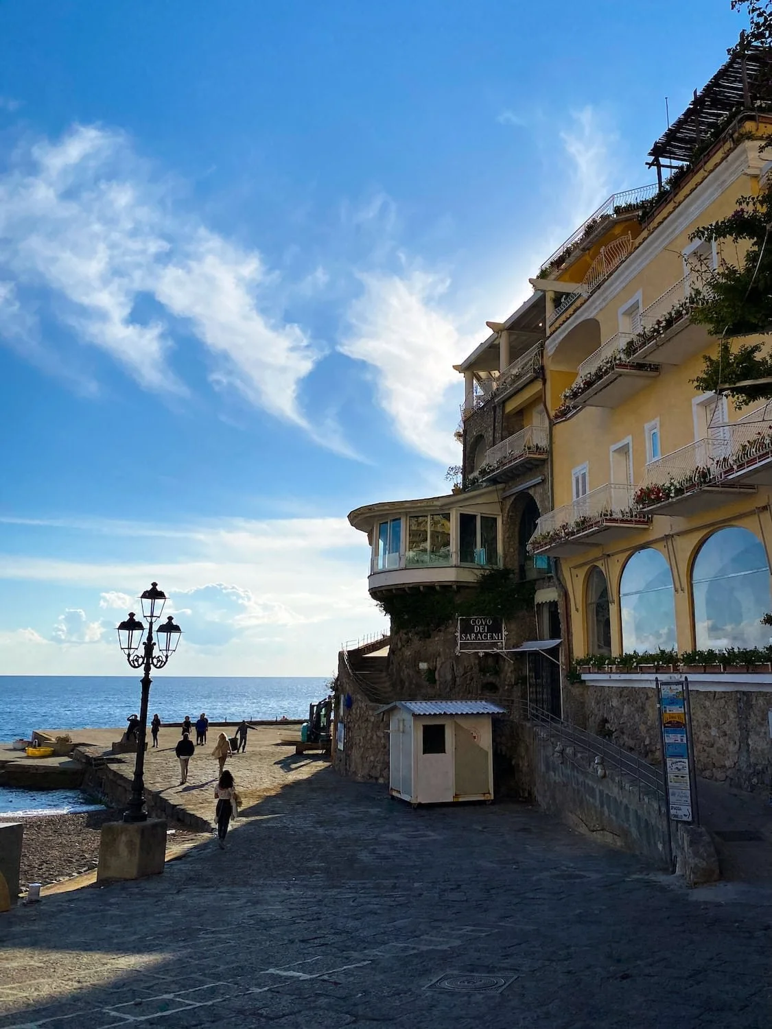 Stone walkway beside the beach in Positano with people strolling toward the sea at sunset. Elegant waterfront buildings and vintage street lamps line the coastal promenade on the Amalfi Coast.