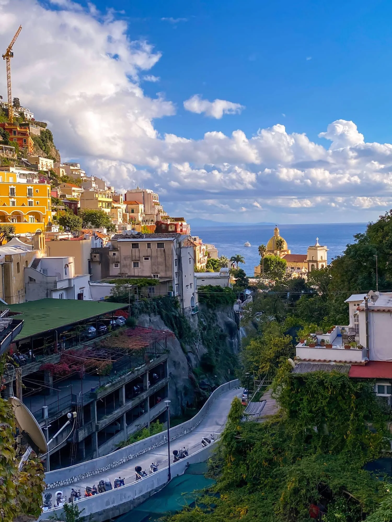 Scenic overlook in Positano featuring winding roads, hillside buildings, and a church dome with the sea in the background. Puffy white clouds stretch across the bright blue sky above the Amalfi Coast.