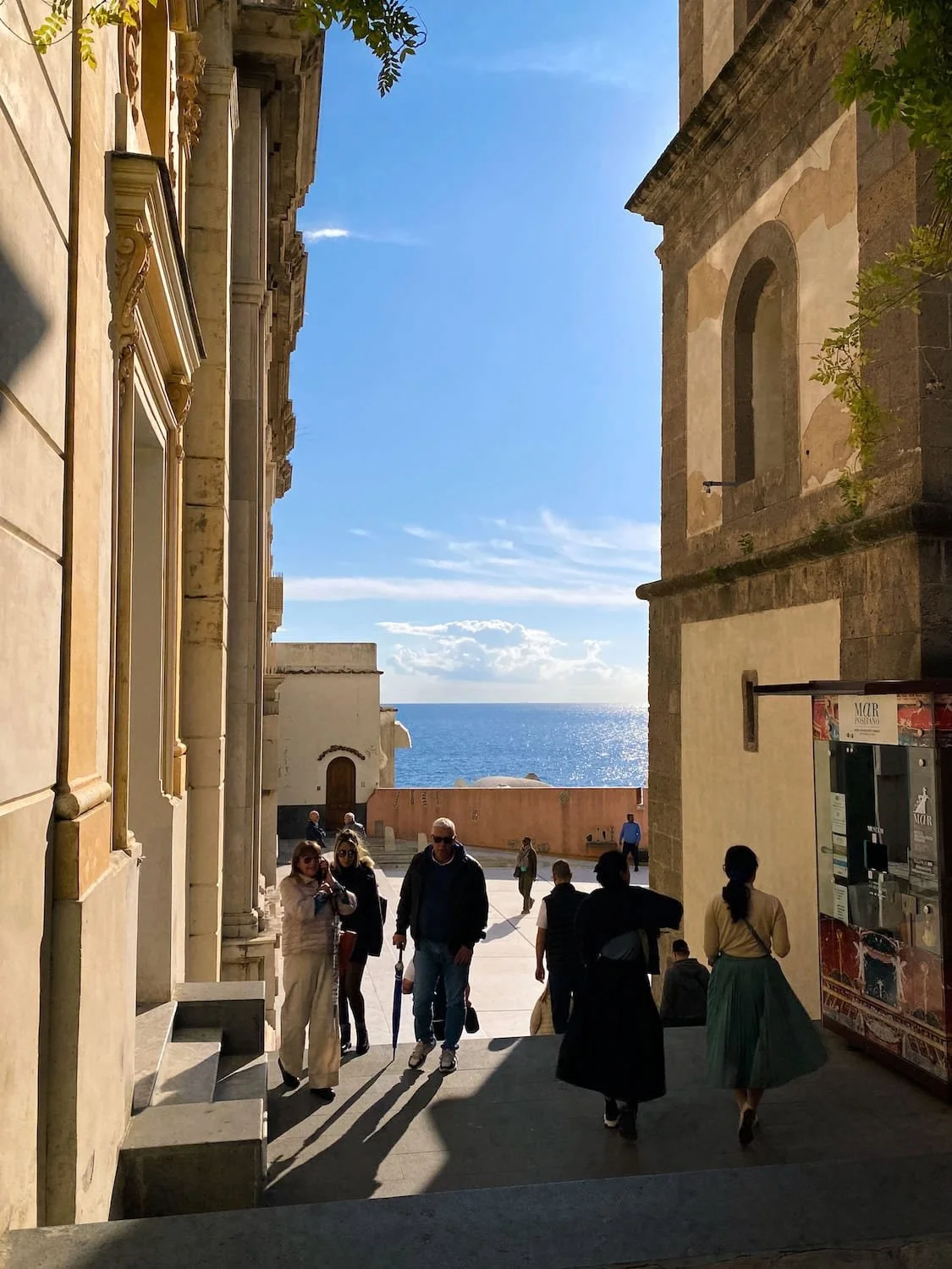Narrow stone walkway in Positano framed by historic buildings and opening to a bright view of the sparkling sea. Visitors stroll through the sunlit passage toward the waterfront.