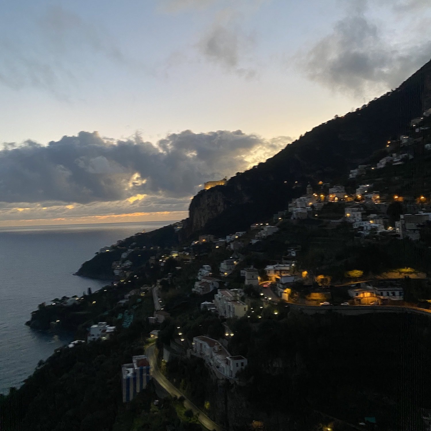 Amalfi Coast at dusk with hillside homes illuminated along winding roads and cliffs above the sea. The dramatic evening view reflects the beauty that shapes where to stay in Amalfi.