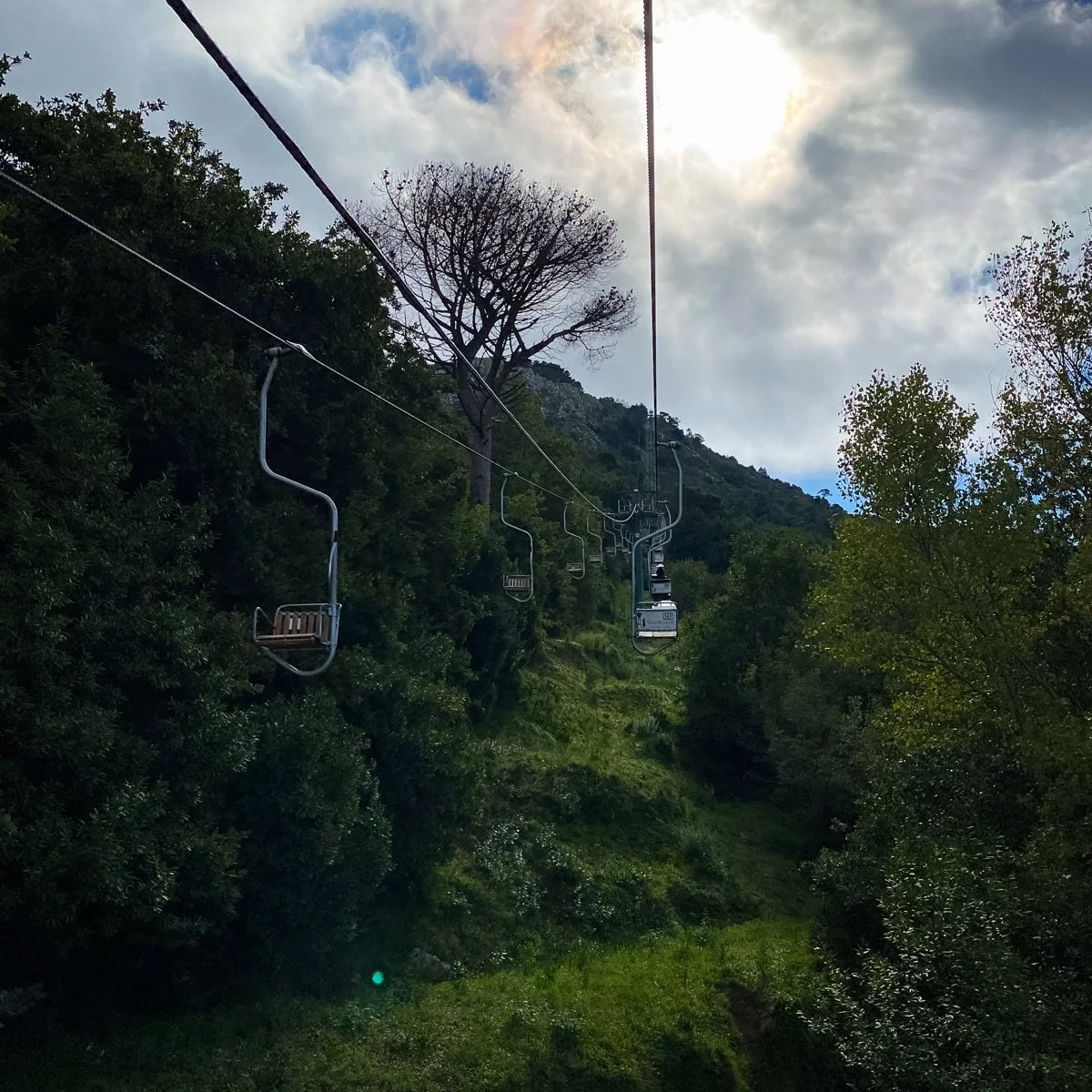 A scenic ride up Monte Solaro on open-air chairlifts, gliding above green forested hills under a partly cloudy sky with sun peeking through — part of the iconic Monte Solaro Chairlift experience.