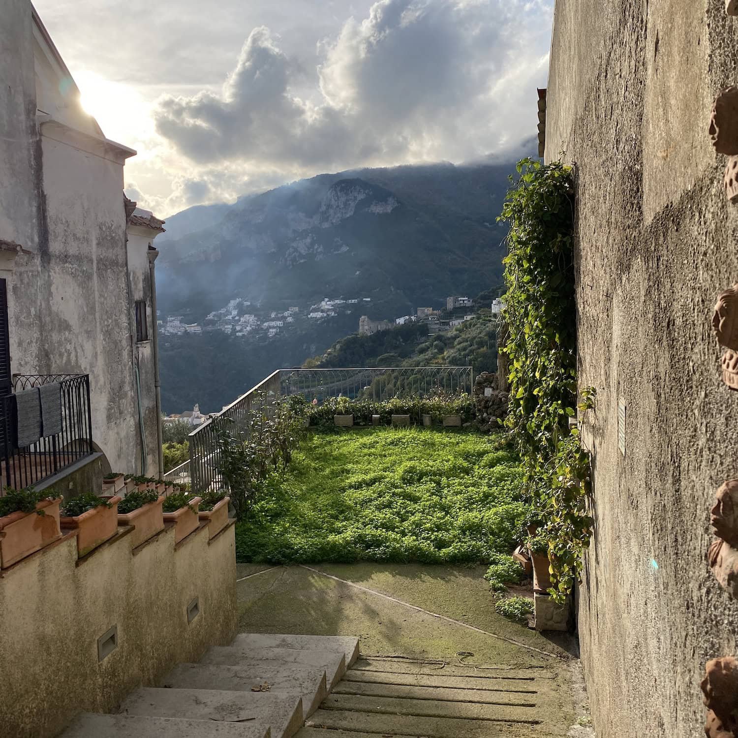 Garden terrace with greenery and mountain views framed by stone walls and sunlight filtering through clouds. The peaceful setting reflects quiet areas to stay in Amalfi away from crowds.