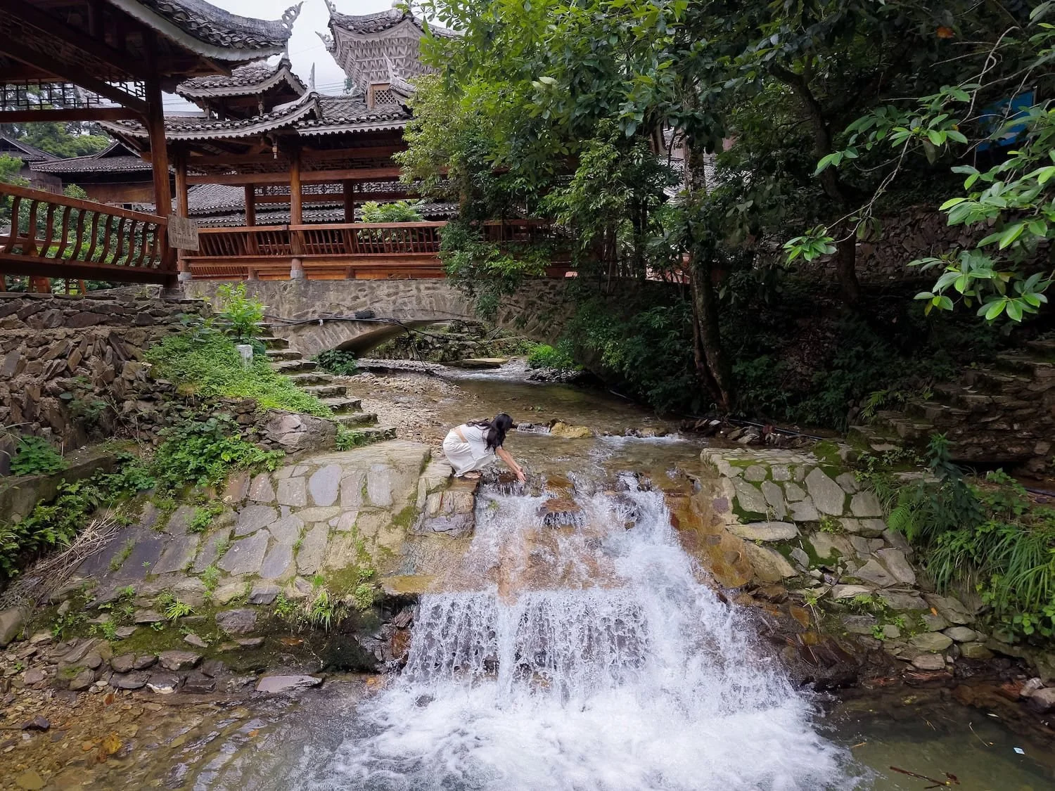A traveler crouches beside a small waterfall beneath a traditional wooden pavilion. A group tour China can help travelers fit multiple scenic and cultural attractions into a limited timeframe.