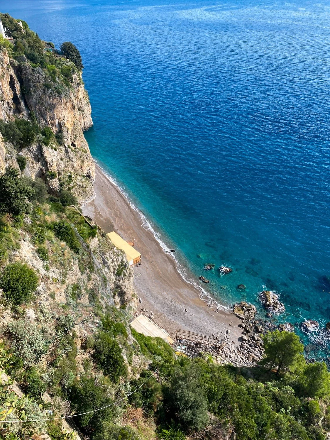 View from above of a quiet beach tucked beneath steep rocky cliffs along the Amalfi Coast near Ravello. Clear turquoise water laps against the shoreline beside rugged coastal scenery.