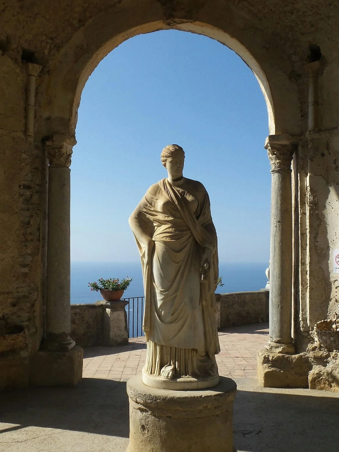 Marble statue framed by a stone archway overlooking the blue Mediterranean Sea in Ravello. Ancient architectural details and coastal views create a dramatic scene at this historic villa.