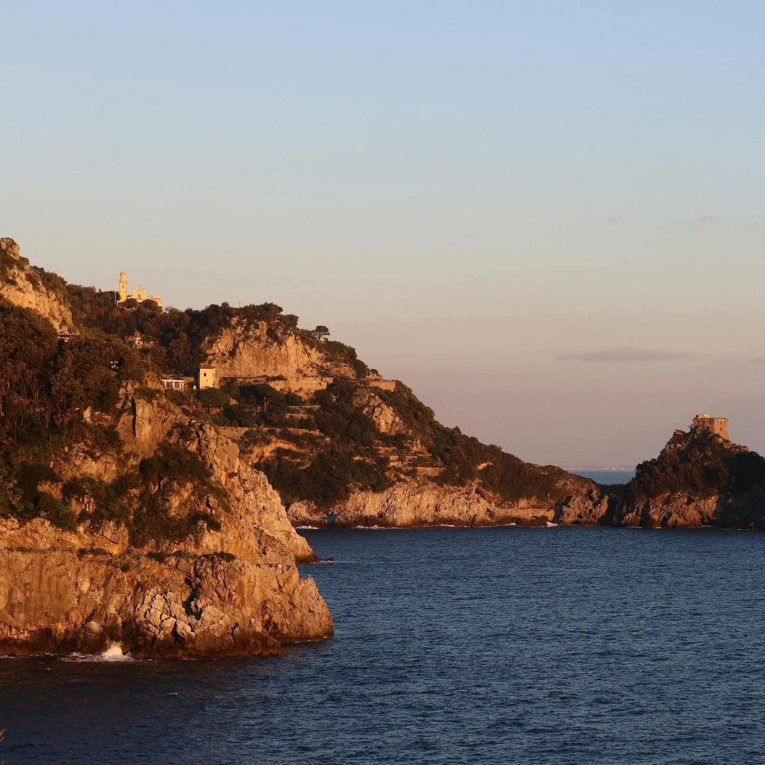 Rocky cliffs glowing in warm sunset light with a small tower perched on a point above the sea along the Amalfi Coast. Watching golden hour along the coastline is one of the peaceful Amalfi Coast things to do.