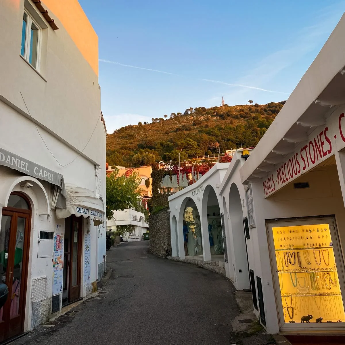 A quaint street in Anacapri featuring jewelry shops and a gelateria near the ticket office for the Monte Solaro Chairlift, captured during golden hour.