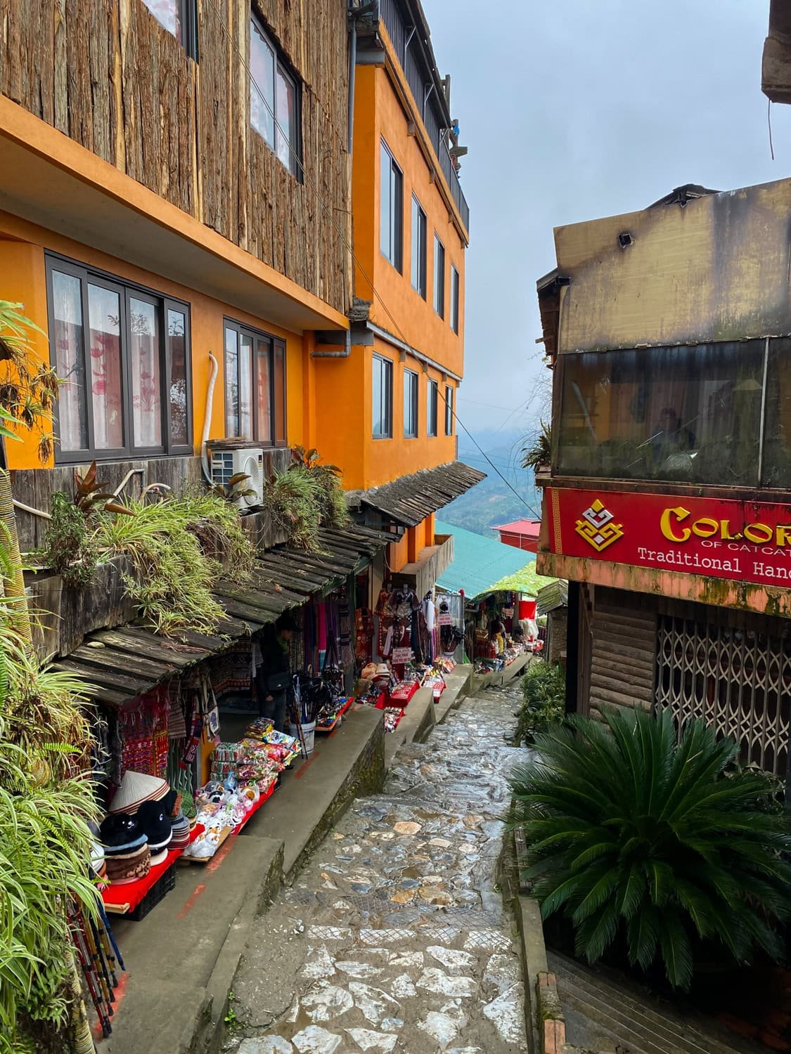 A quiet alleyway in Cat Cat Village with local shops on both sides, vibrant crafts on display, and an orange building contrasting against the cloudy mountain background.