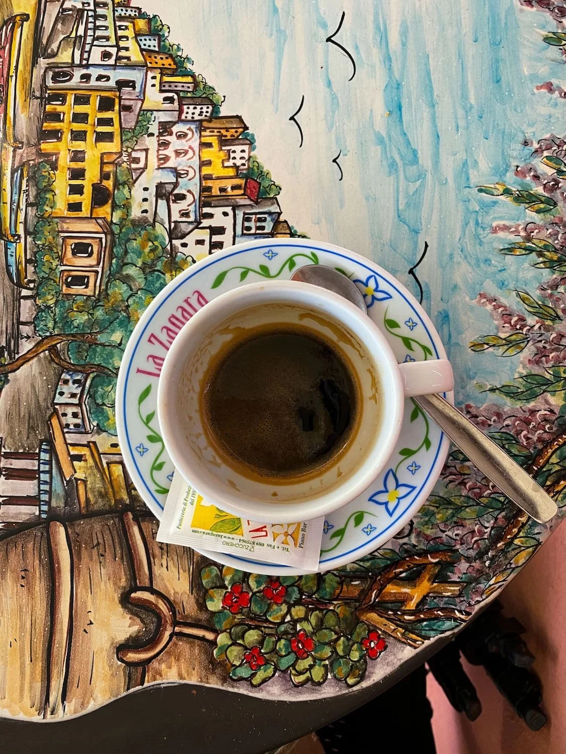 Small espresso cup on a hand painted ceramic table featuring a colorful Positano coastal scene. A silver spoon and sugar packet rest beside the coffee during a relaxing café break on the Amalfi Coast.