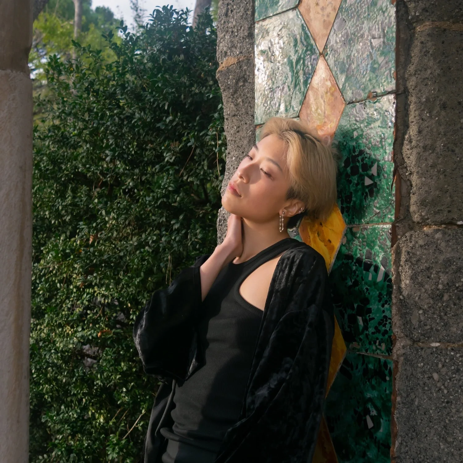 Traveler with short blonde hair leaning against a colorful tiled wall surrounded by greenery in a quiet Amalfi Coast setting highlighted in this Amalfi travel guide. Warm sunlight and textured stone columns frame a relaxed portrait moment.