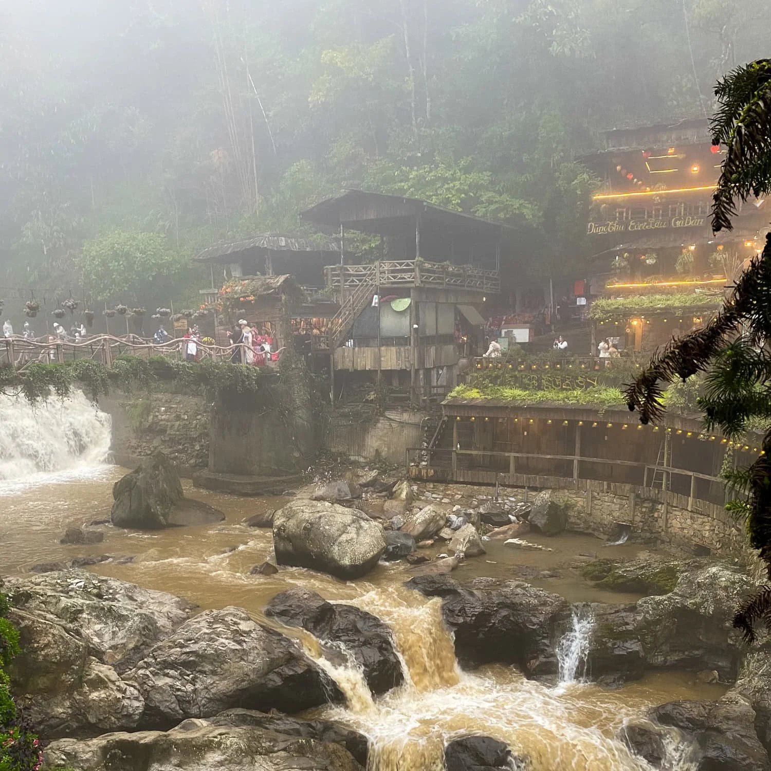 A foggy riverside view in Cat Cat Village showing a cascading stream flowing between rocky banks, with charming wooden buildings lit up on the hillside.