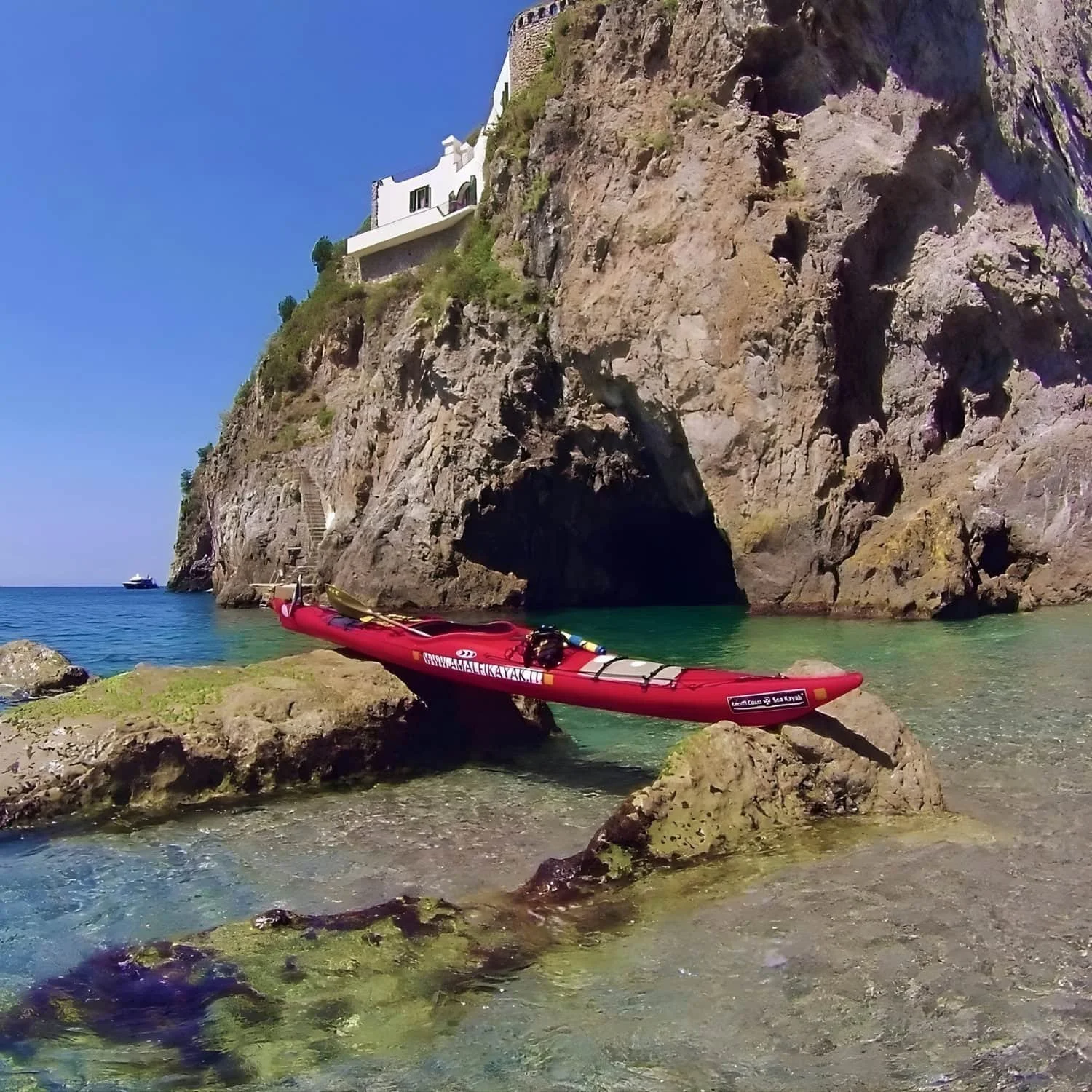 Red kayak resting on rocks beside clear turquoise water and a sea cave beneath steep coastal cliffs. Exploring hidden coves by kayak is one of the adventurous Amalfi Coast things to do.
