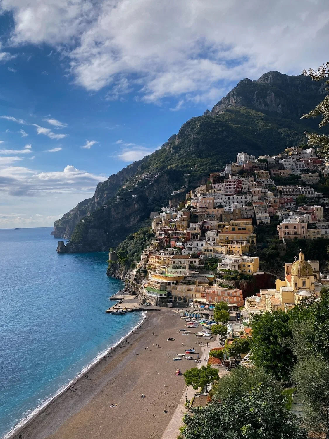 Panoramic view of Positano beach and colorful cliffside homes beside the turquoise Mediterranean Sea. Mountain peaks rise above the town under a bright blue sky with scattered clouds.