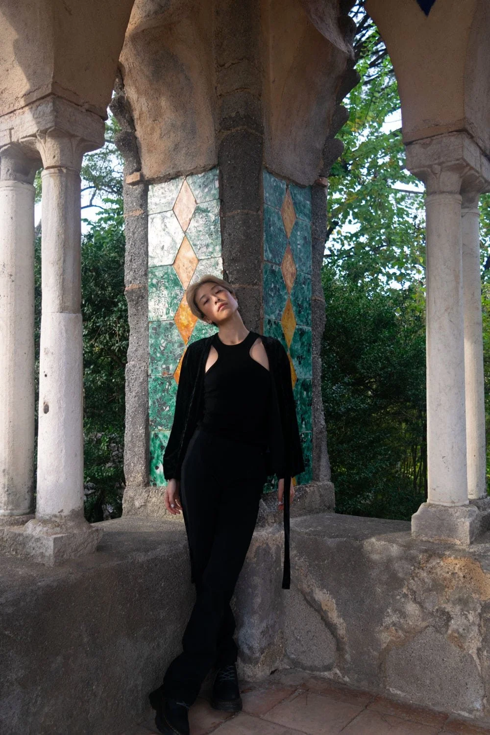 Woman dressed in black standing beneath a tiled stone pavilion with arched columns and green mosaic details in Ravello. The historic structure and shaded garden setting highlight one of the unique things to do in Ravello along the Amalfi Coast.