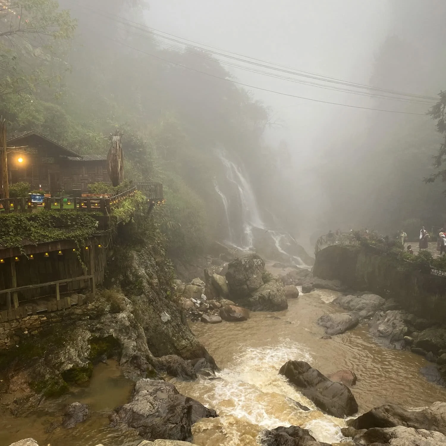 A foggy landscape in Cat Cat Village featuring a dramatic waterfall cascading into a rocky stream, with small wooden buildings clinging to the cliffs nearby.