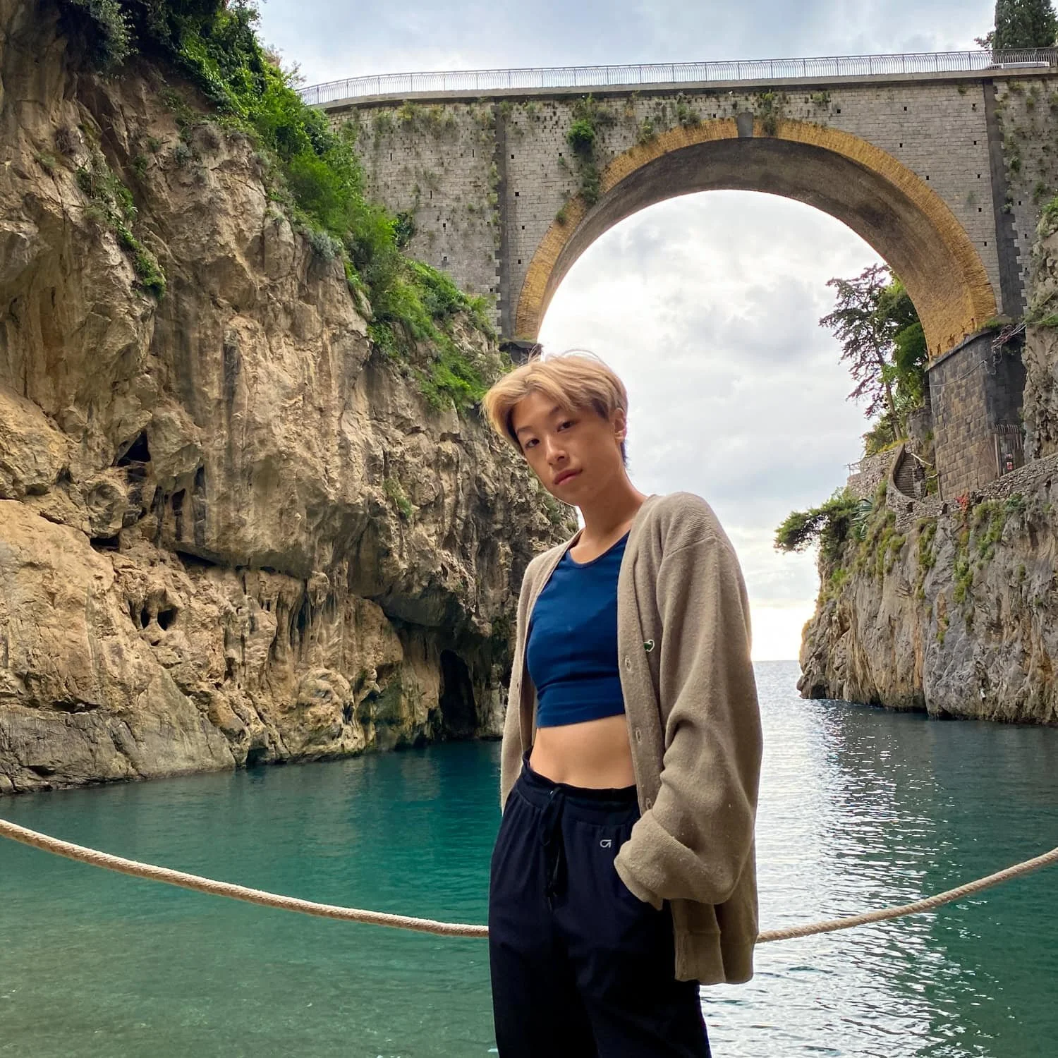 Traveler standing beside turquoise water beneath the tall stone arch bridge at Fiordo di Furore on the Amalfi Coast. Visiting this dramatic coastal gorge is one of the scenic Amalfi Coast things to do.