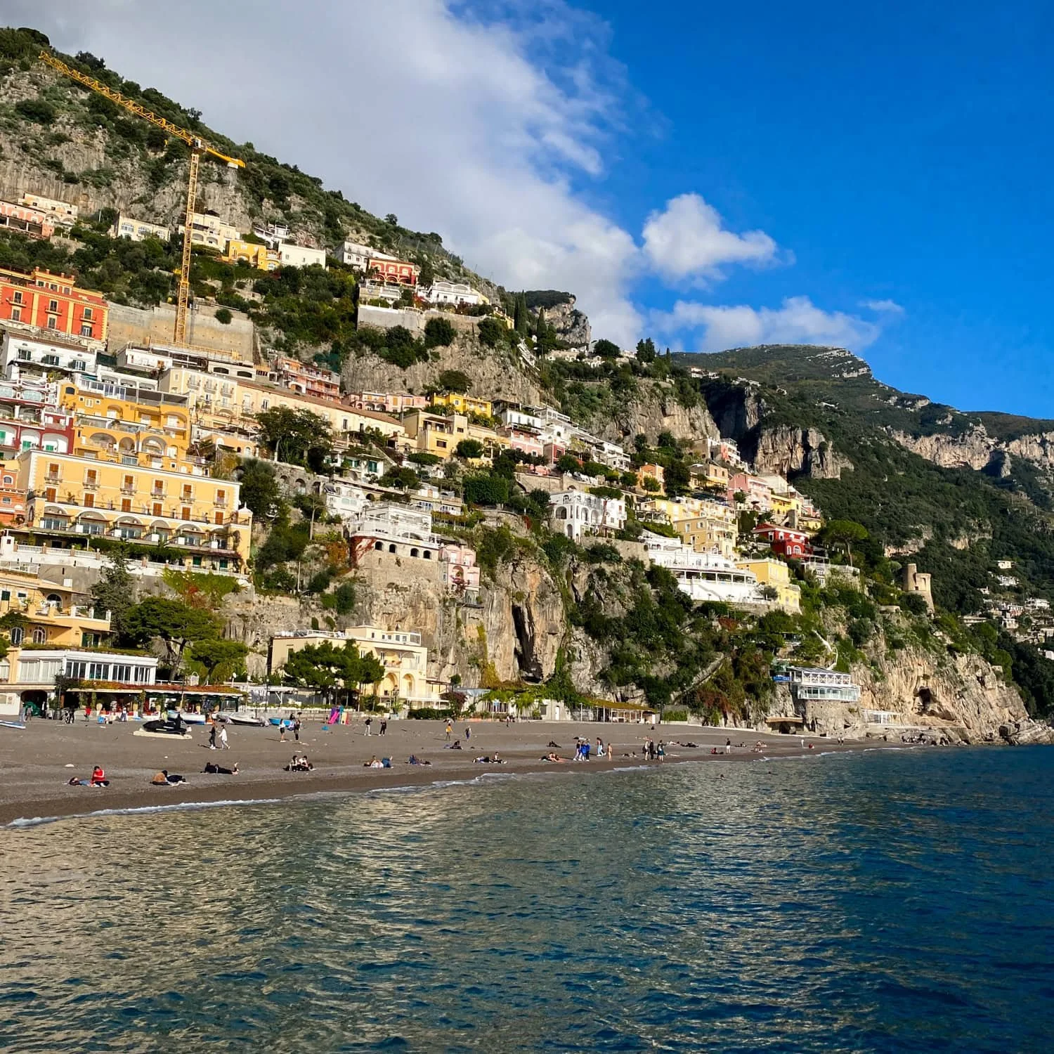 Colorful cliffside buildings tower above a sandy beach and calm sea along the Amalfi Coast in this Amalfi travel guide. People relax near the shoreline while steep rock formations frame the vibrant seaside village.