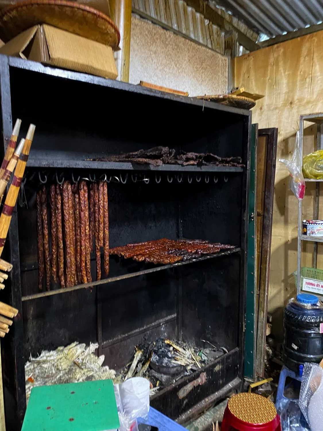 A rustic smoker in Cat Cat Village filled with hanging sausages and grilled meat, with firewood burning beneath, showing traditional food preparation methods.