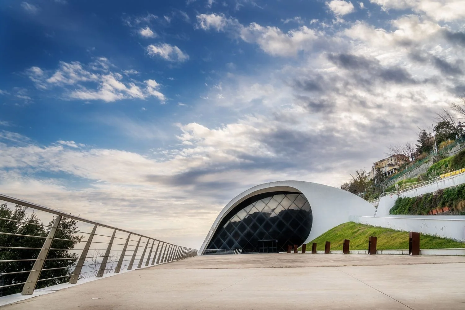 Modern curved white architecture of the Oscar Niemeyer Auditorium in Ravello beneath a dramatic cloudy sky. The sleek concert hall contrasts with the surrounding hillside landscape on the Amalfi Coast.