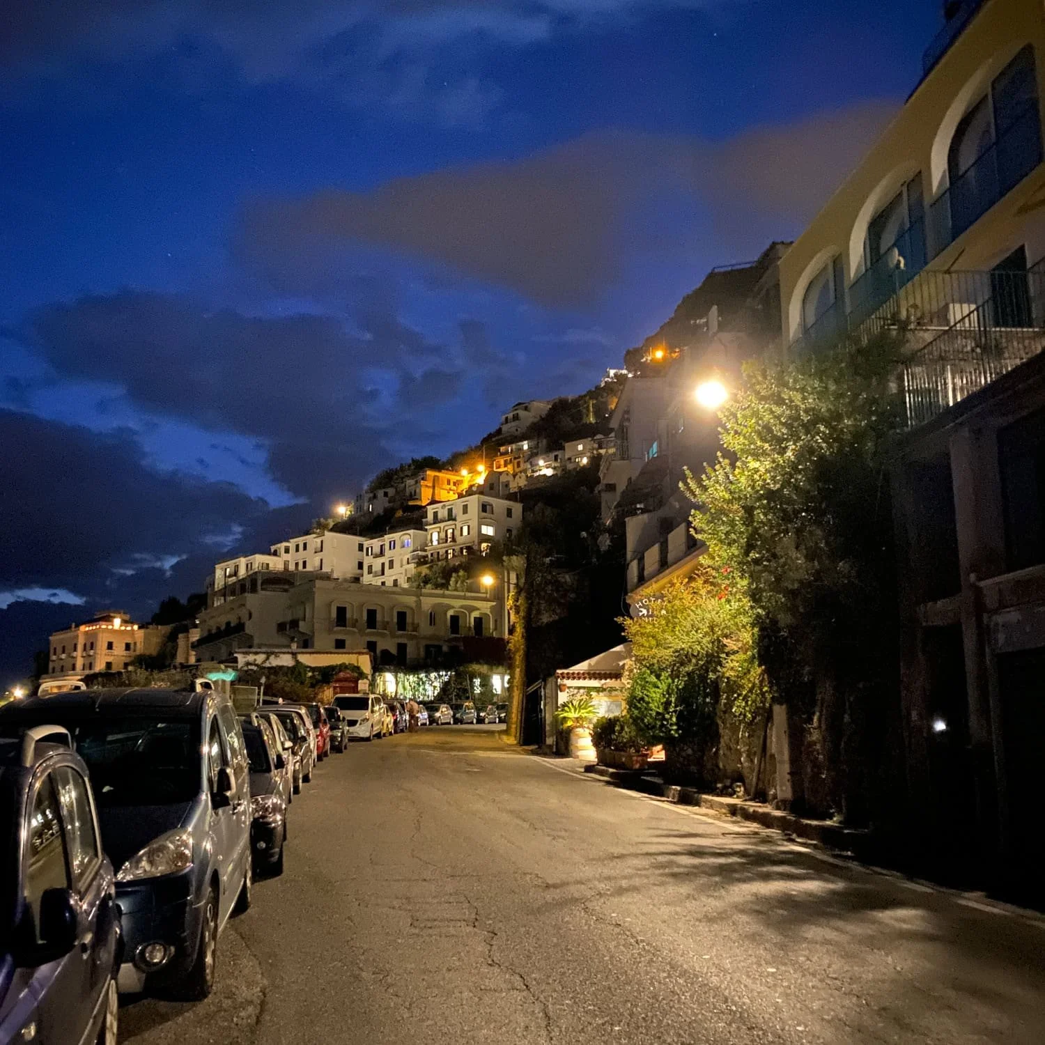 Quiet Amalfi Coast street at night with parked cars glowing streetlights and hillside homes illuminated against a deep blue sky in this Amalfi travel guide. The winding road and warm window lights create a peaceful evening atmosphere.