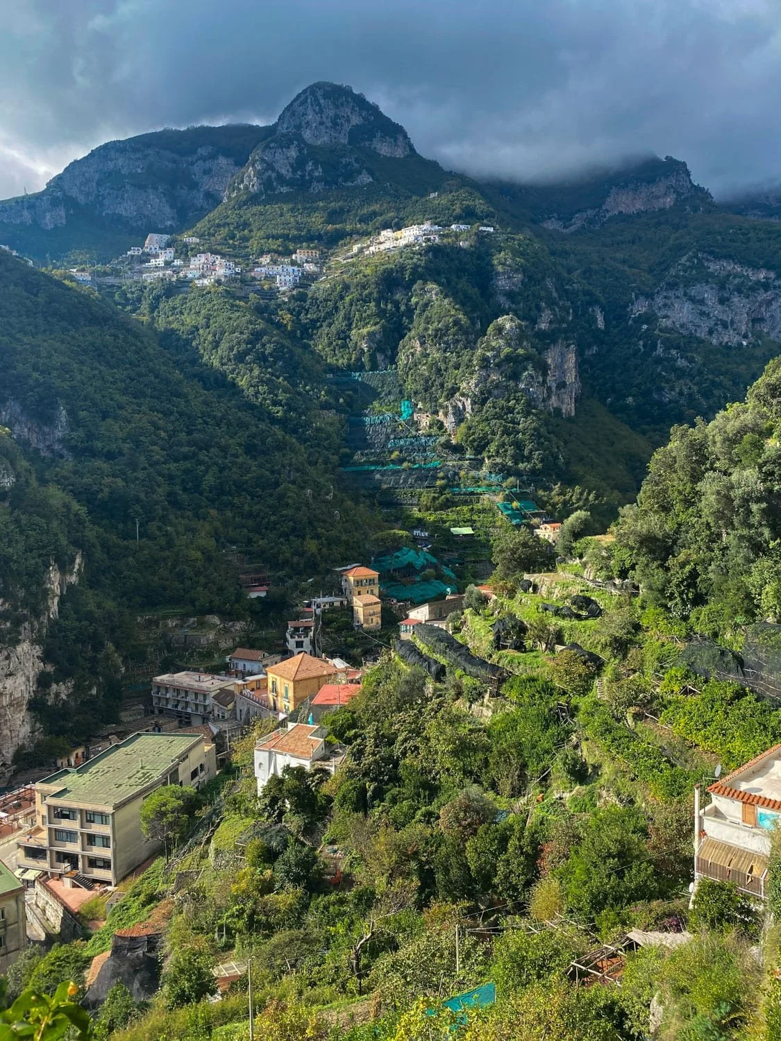 Mountain village surrounded by lush green hills and dramatic cliffs near Ravello on the Amalfi Coast. Sunlight breaks through dark clouds over terraced landscapes and clustered hillside homes.