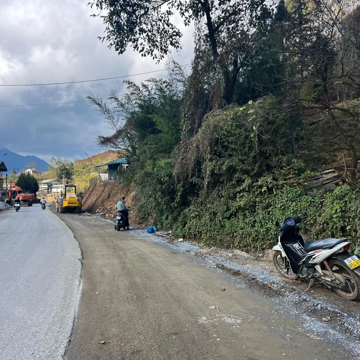 Motorbikes and road construction equipment line a partly paved road in rural Sapa, surrounded by dense greenery and hills in the background. Potential safety risks if you’re visiting Sapa!