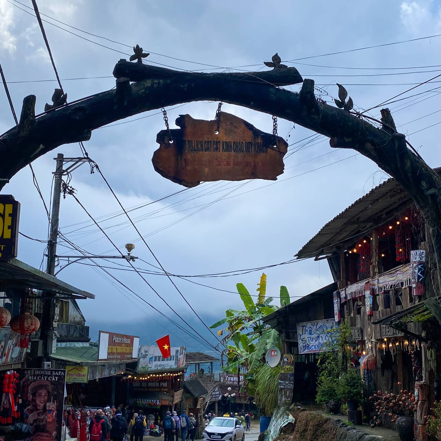 A rustic wooden sign reading “Du Lịch Cát Cát” hangs above the entrance to Cat Cat Village’s tourist area, with shops, people, and a cloudy mountain backdrop visible below.