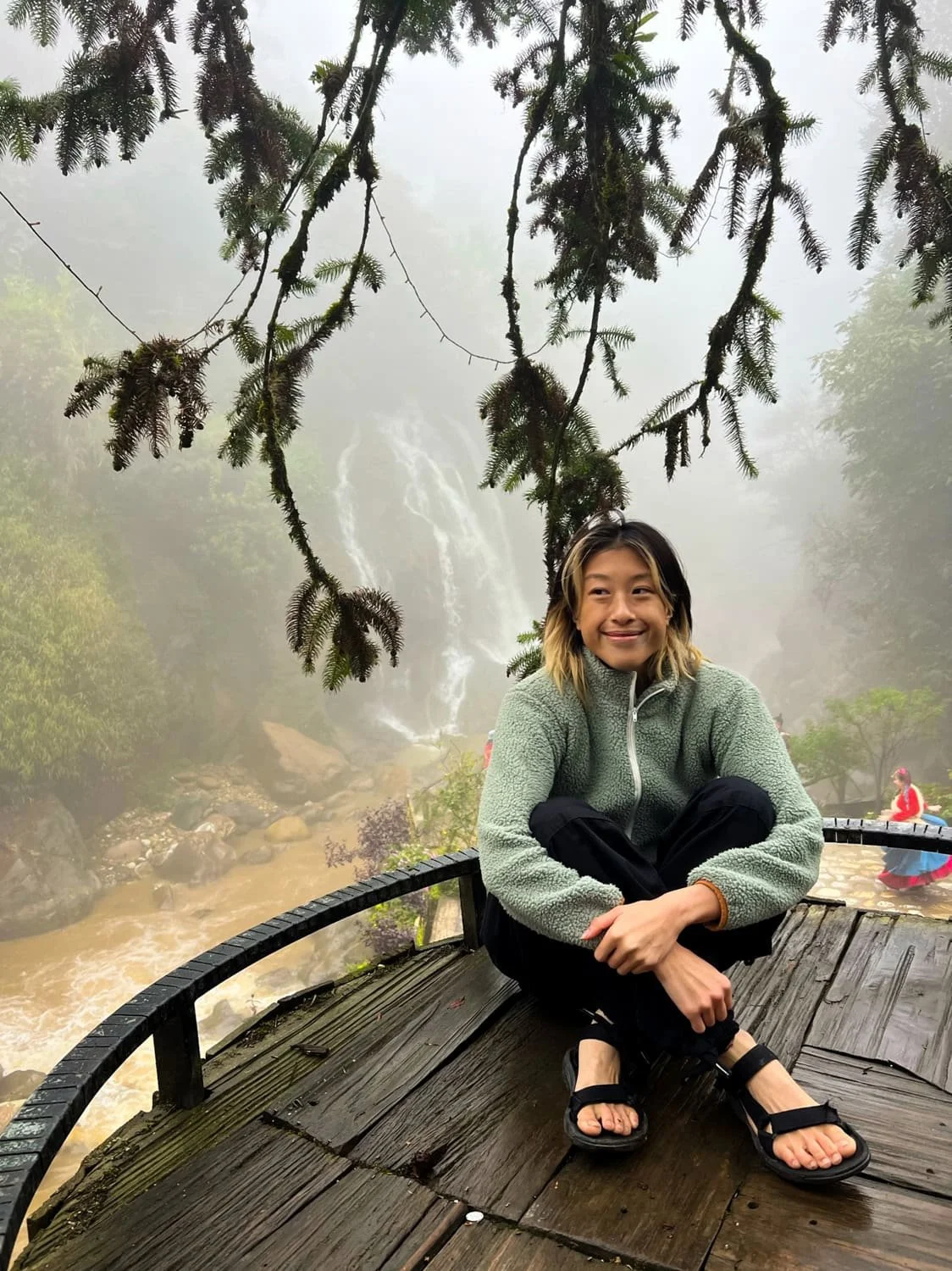 A woman sits cross-legged on a wooden deck under moss-covered tree branches, smiling with the waterfall of Cat Cat Village faintly visible in the mist behind her.