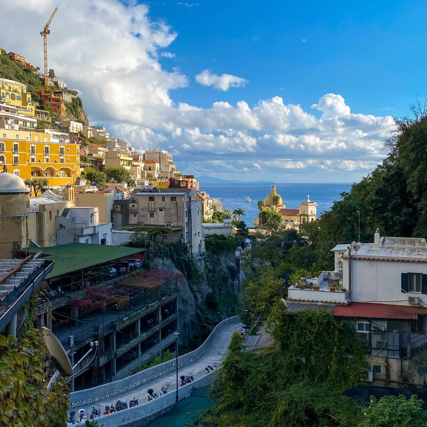 Panoramic view of Positano with pastel buildings cascading down steep cliffs toward the turquoise sea as seen in this Amalfi travel guide. A winding road and domed church overlook the coastline beneath dramatic clouds.
