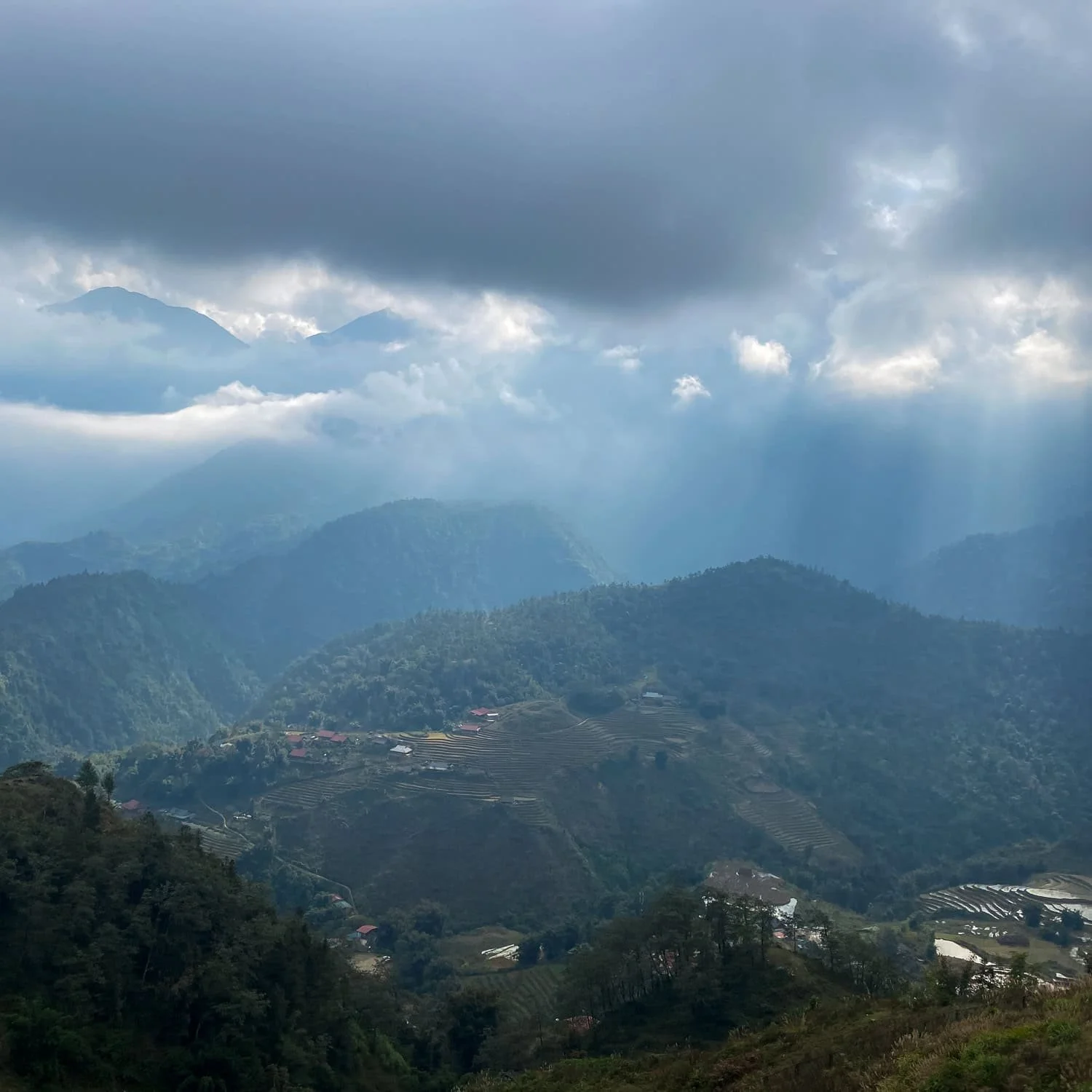 A wide landscape of the Hoàng Liên Son mountain range near Fansipan, dotted with rice terraces and villages under a misty sky.