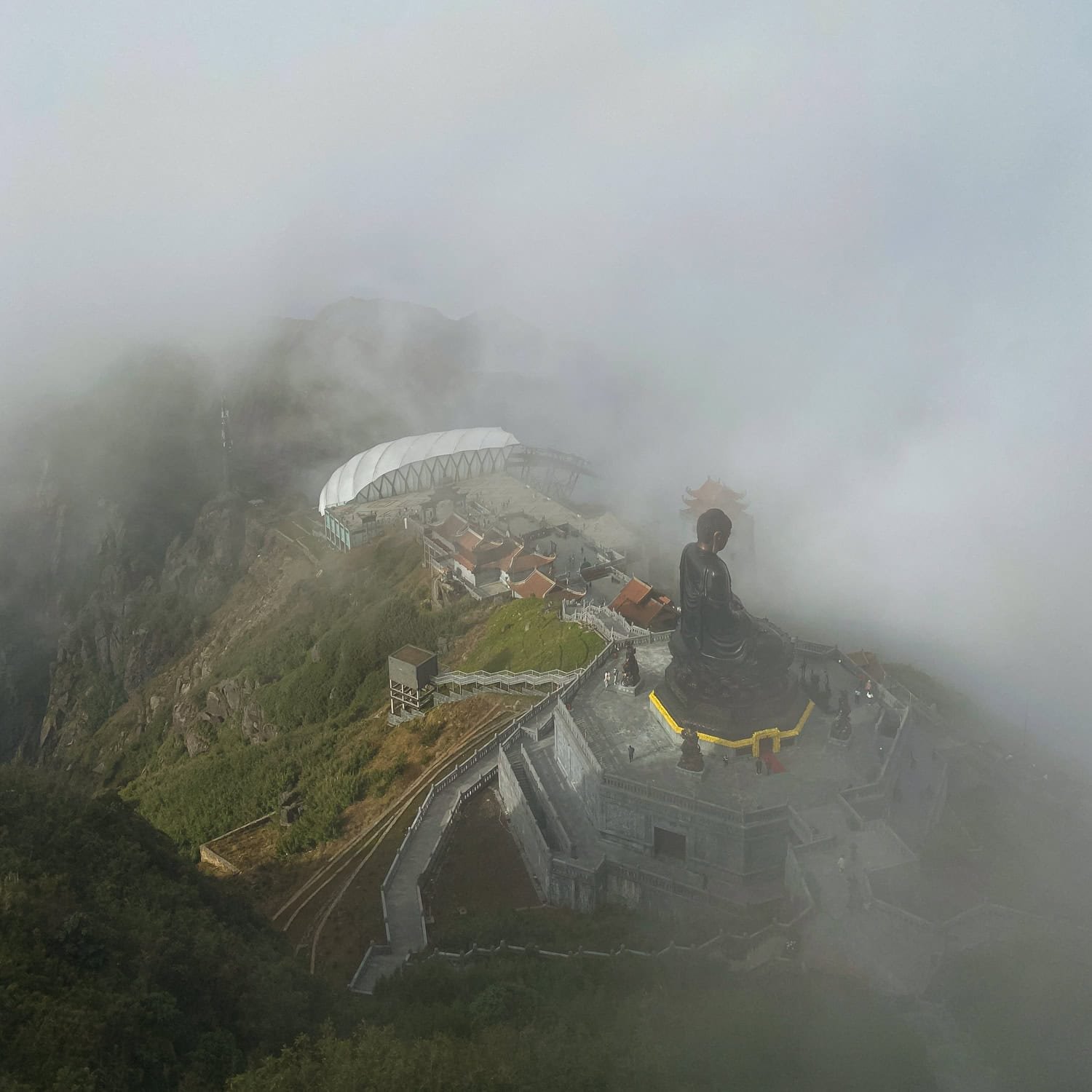 A dramatic aerial view of the Great Buddha statue on Fansipan Mountain, partially covered in clouds, with temples and paths visible below near the Fansipan cable car summit station.