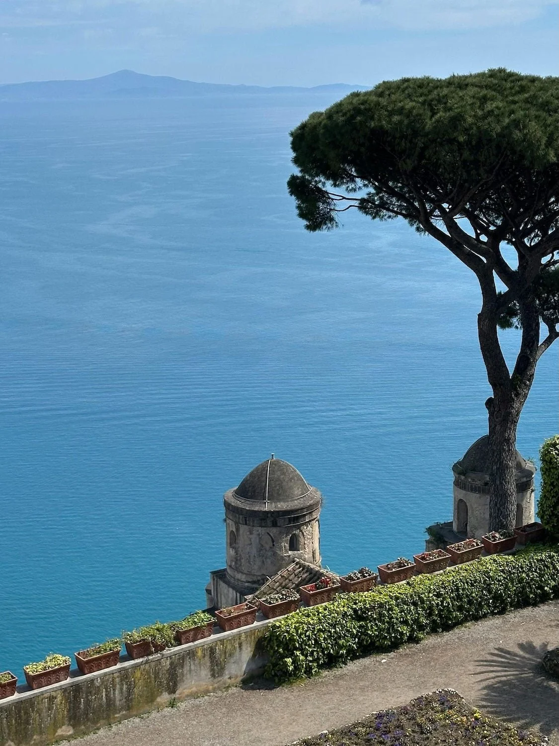 Scenic coastal overlook in Ravello with a tall pine tree, stone towers, and sweeping views of the calm blue sea. Flower-lined terraces frame the dramatic Amalfi Coast landscape below.