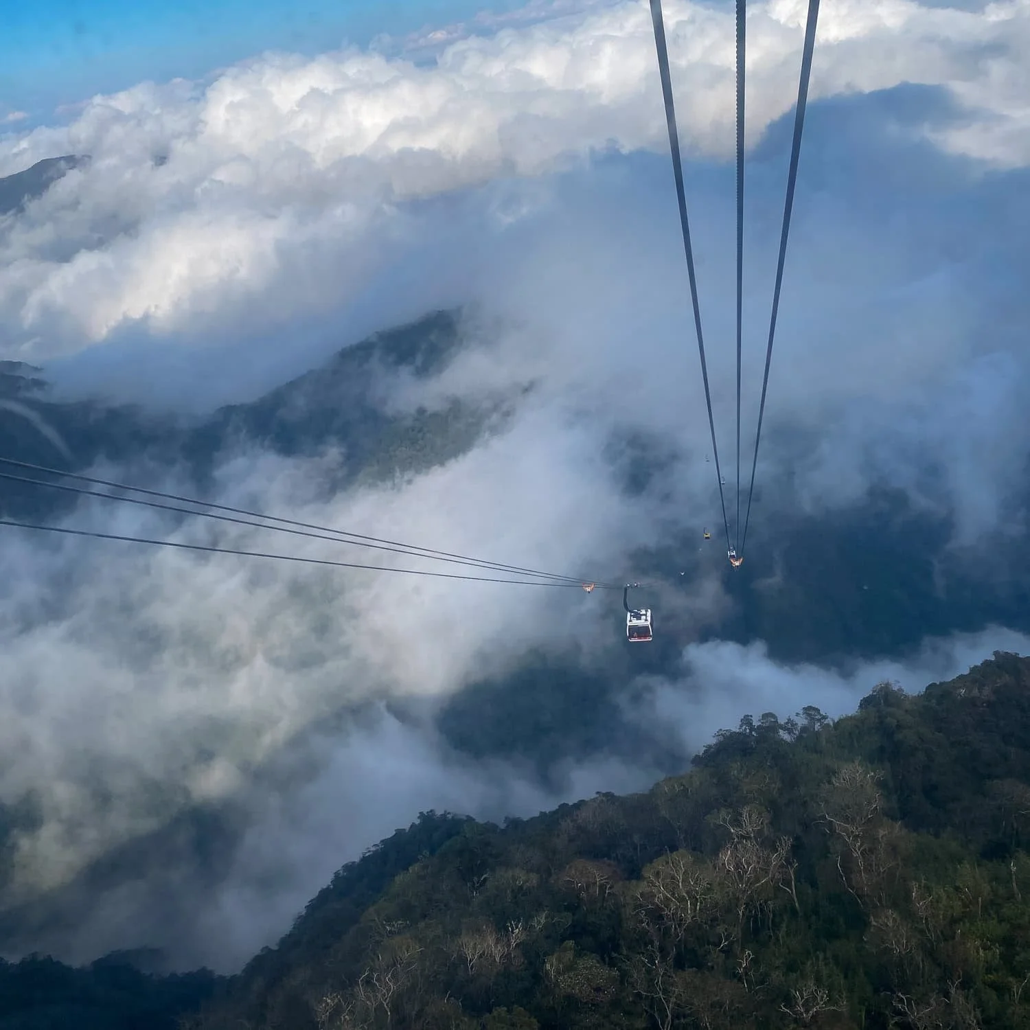 A high-angle view of the Fansipan cable car gliding above thick clouds and forested mountains, suspended between tall peaks in northern Vietnam.