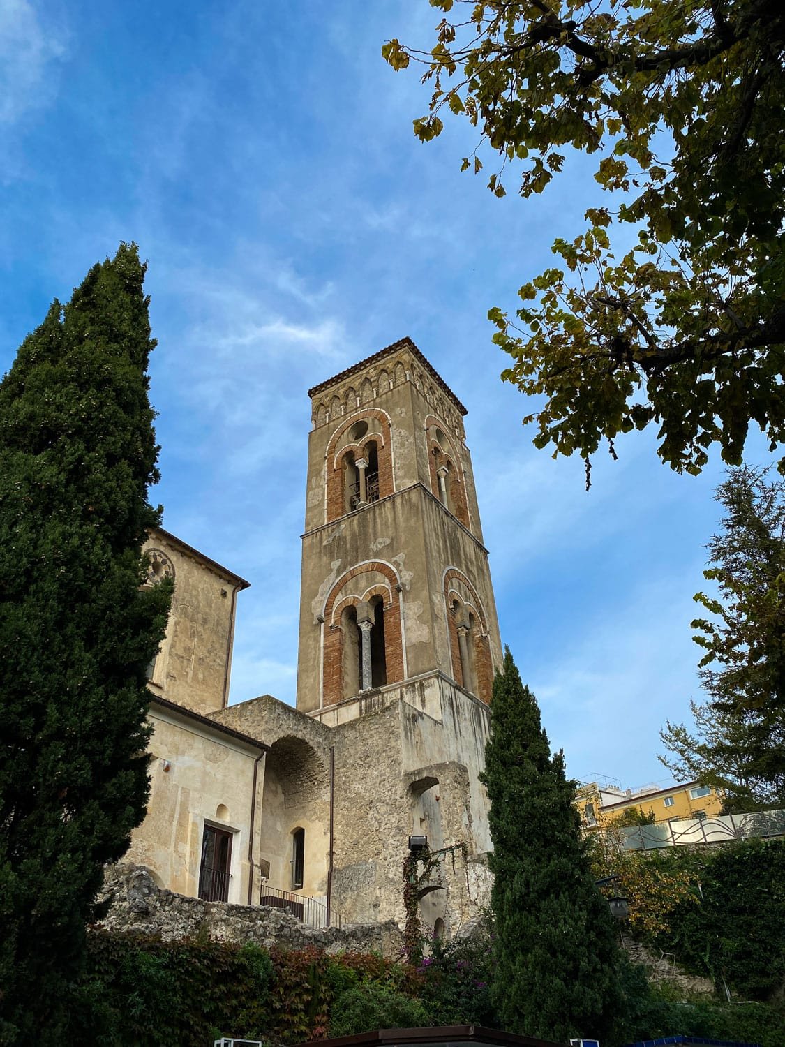 Historic church bell tower in Ravello rising above stone walls and cypress trees under a bright blue sky. Romanesque arches and weathered architecture showcase the town’s historic charm.