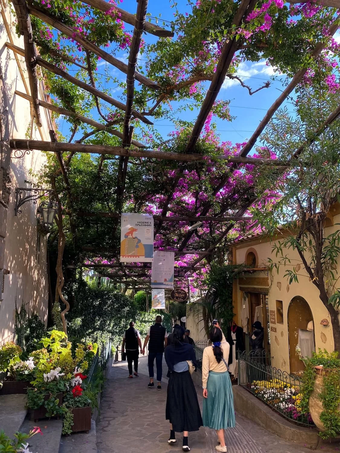 Pedestrian walkway in Positano covered with blooming pink bougainvillea vines overhead. Visitors walk beneath the shaded pergola lined with flowers and small local shops.