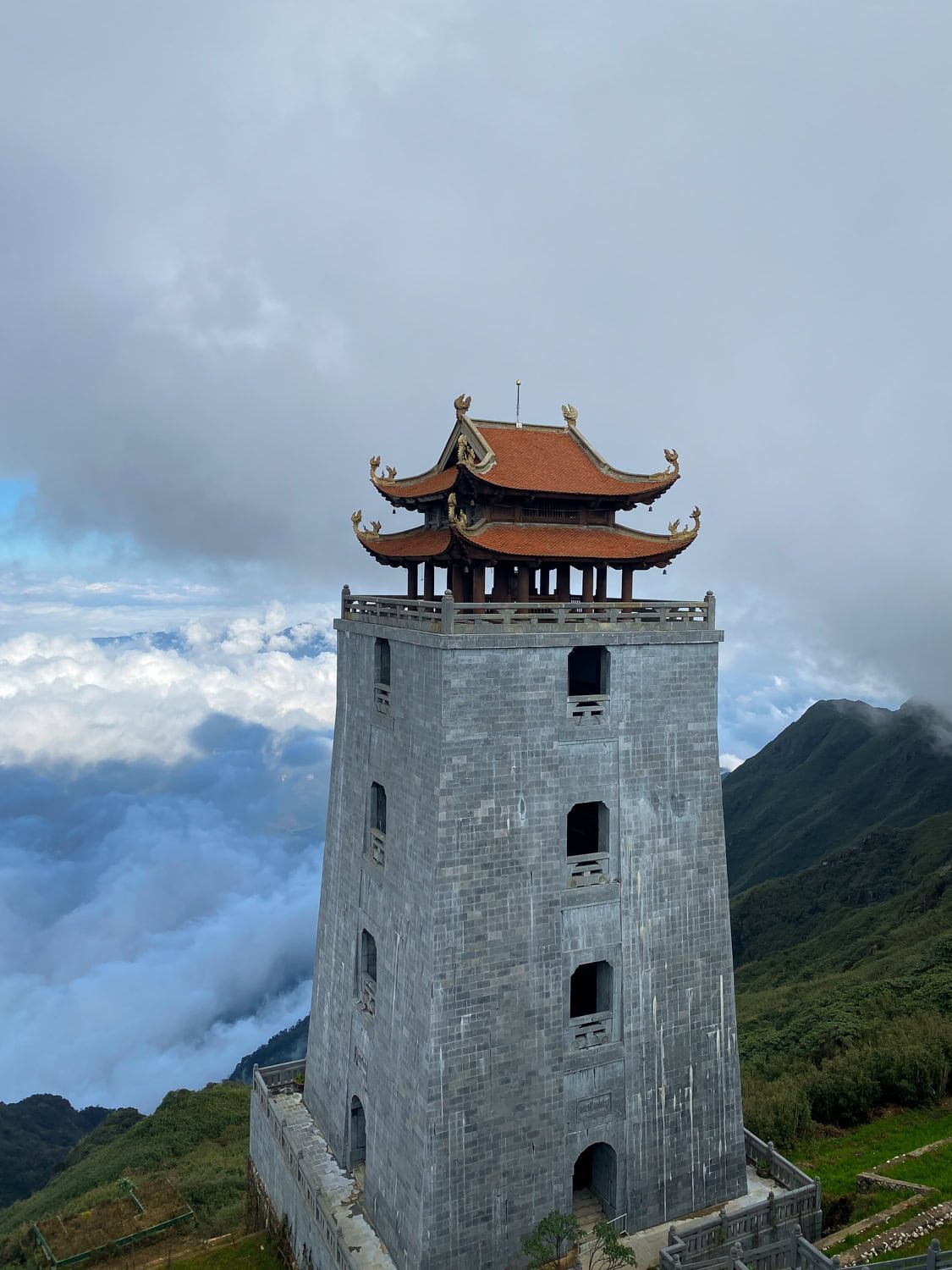 A tall stone pagoda tower with a red-tiled roof sits on a high ridge near the summit of Fansipan, surrounded by clouds and rugged peaks.