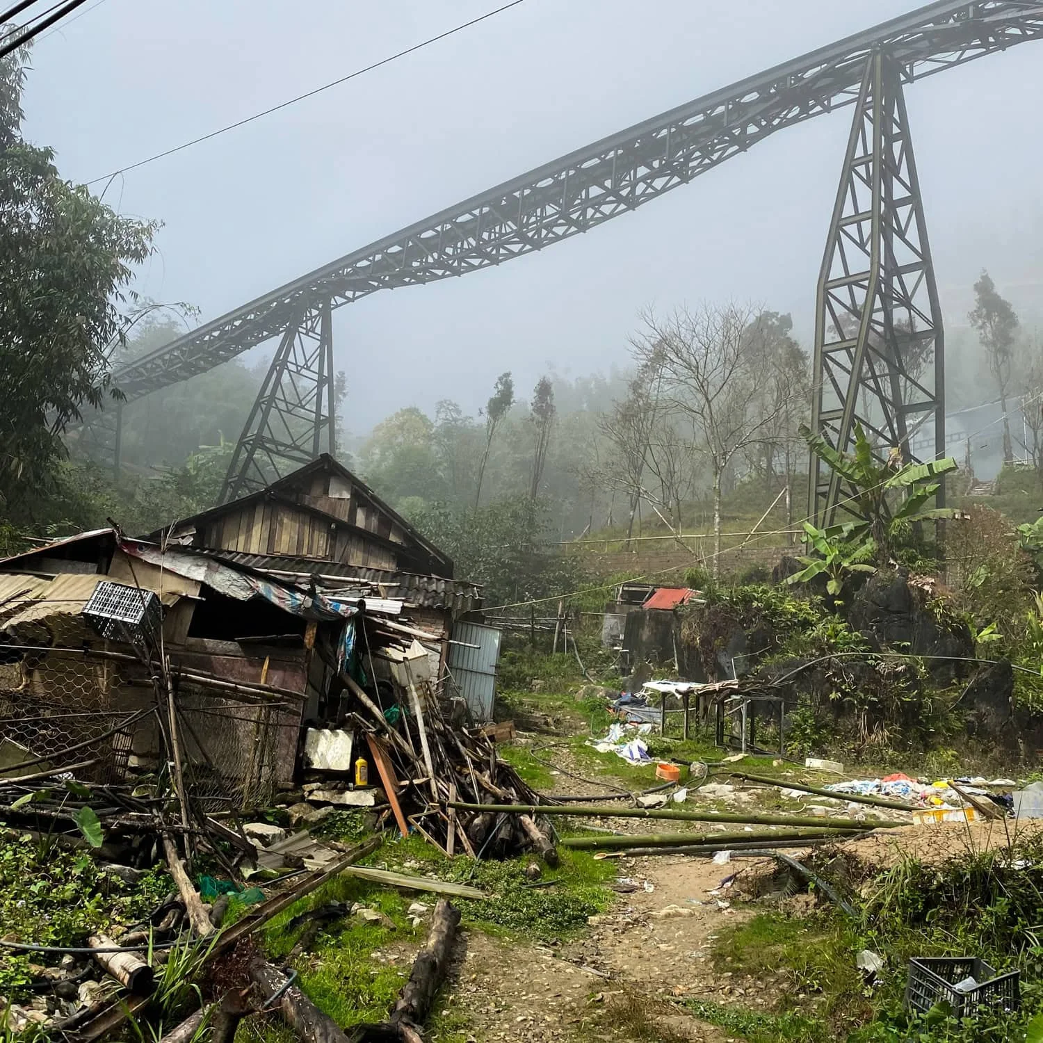A misty scene in Cat Cat Village featuring an old wooden house surrounded by debris and greenery, with a towering bridge structure cutting across the foggy sky.