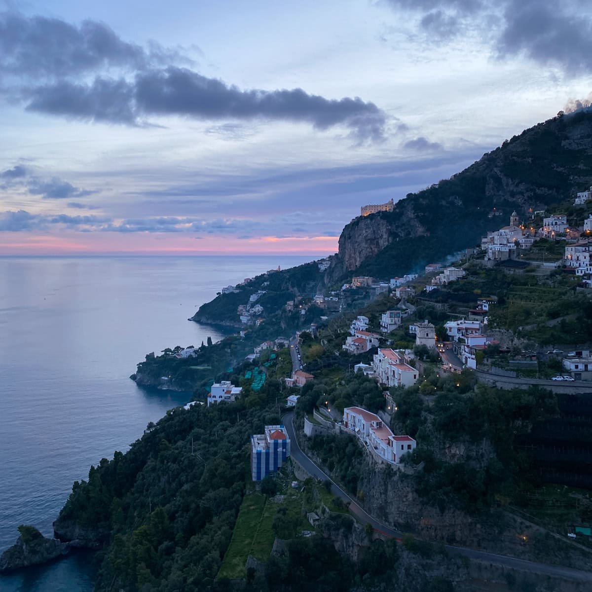 Twilight descends on a serene coastal village along the Amalfi Coast, with lights beginning to glow from cliffside homes symbolizing the final reflections on choosing between Amalfi or Positano.