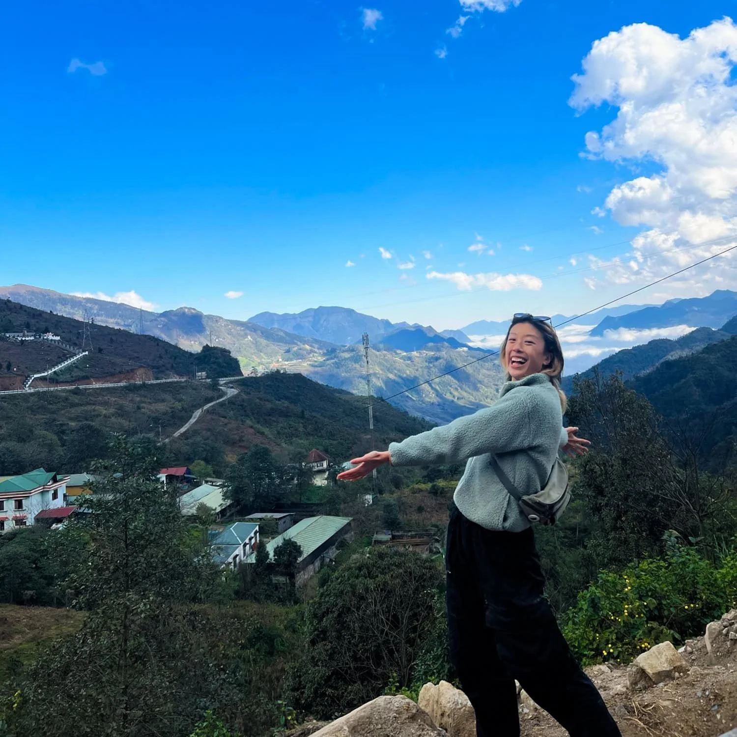 A woman poses joyfully with arms outstretched on a roadside overlooking the scenic, mountainous landscape of Cat Cat Village under a bright blue sky.