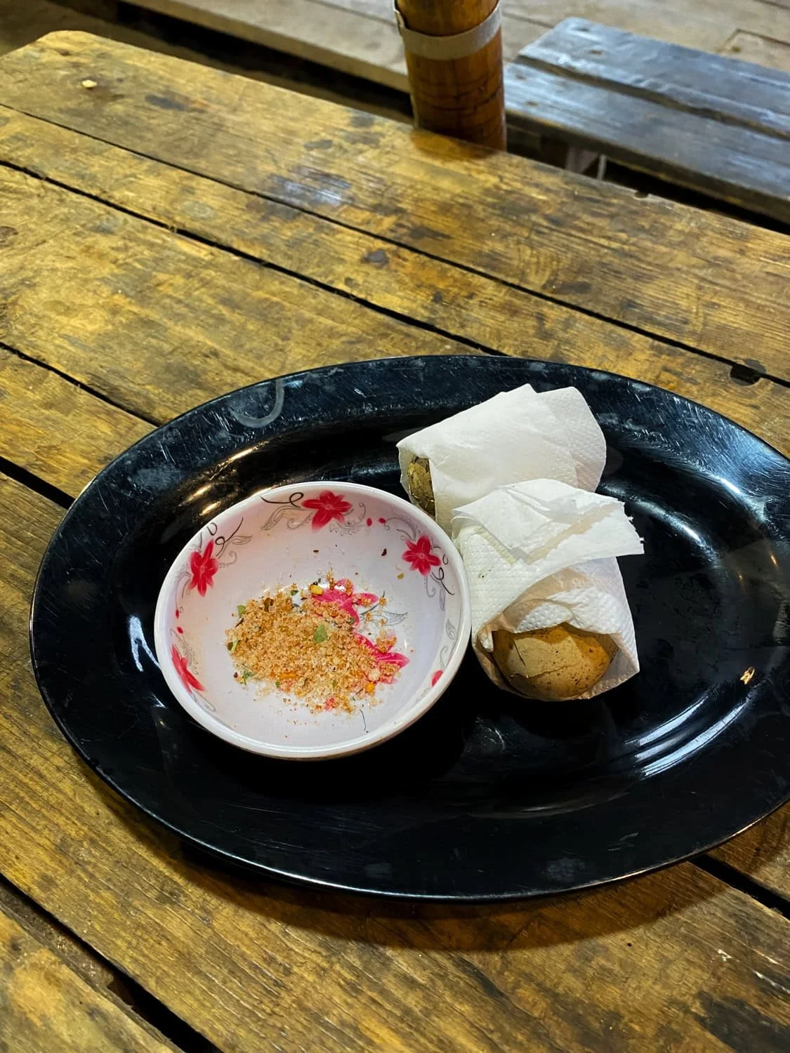 A black plate holds two grilled eggs wrapped in napkins and a small bowl of colorful salt and spice mix, served on a rustic wooden table in Cat Cat Village.