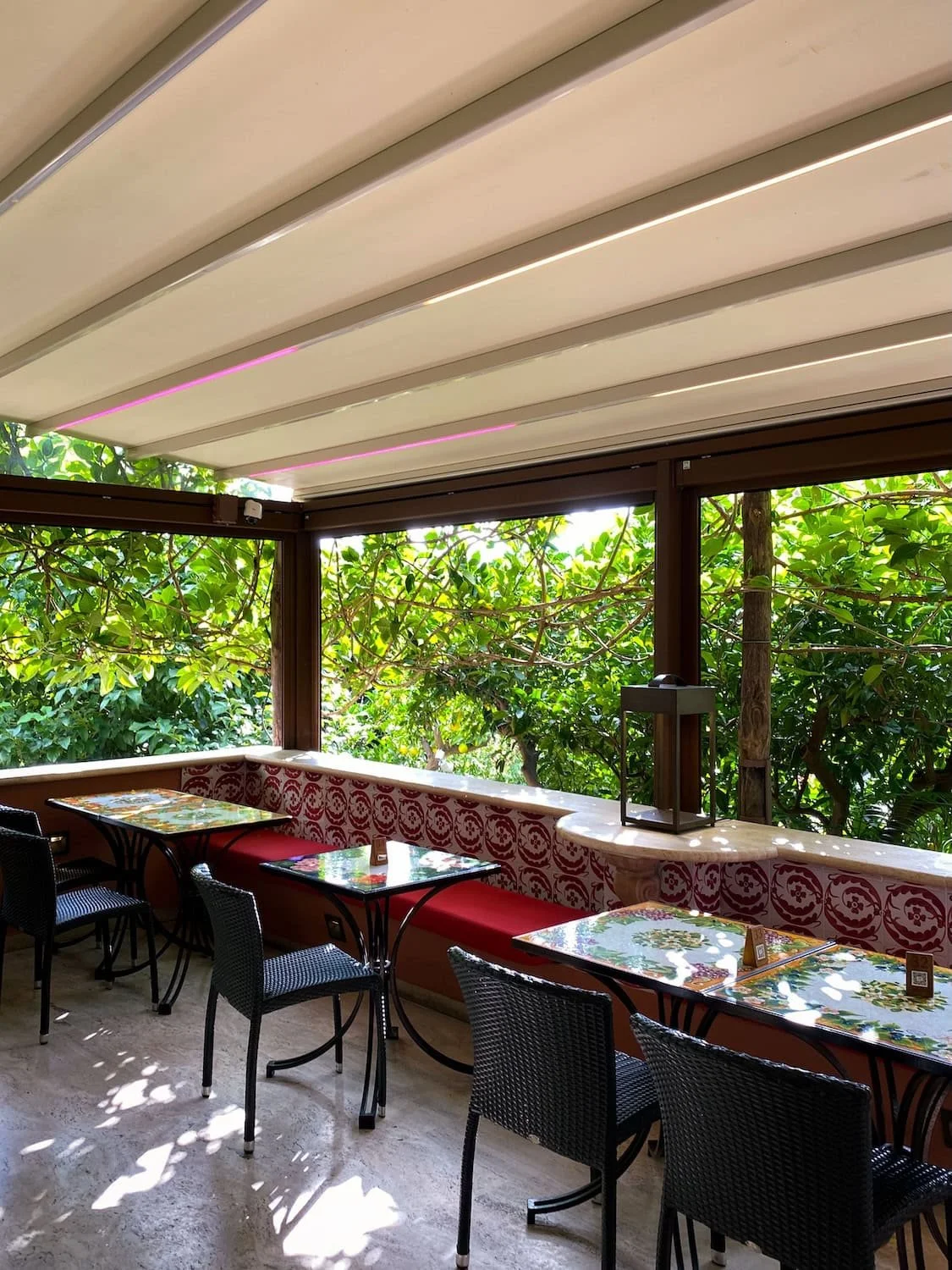 Outdoor café seating in Positano surrounded by lush green trees and filtered sunlight. Patterned tables and red cushioned benches create a peaceful terrace atmosphere.