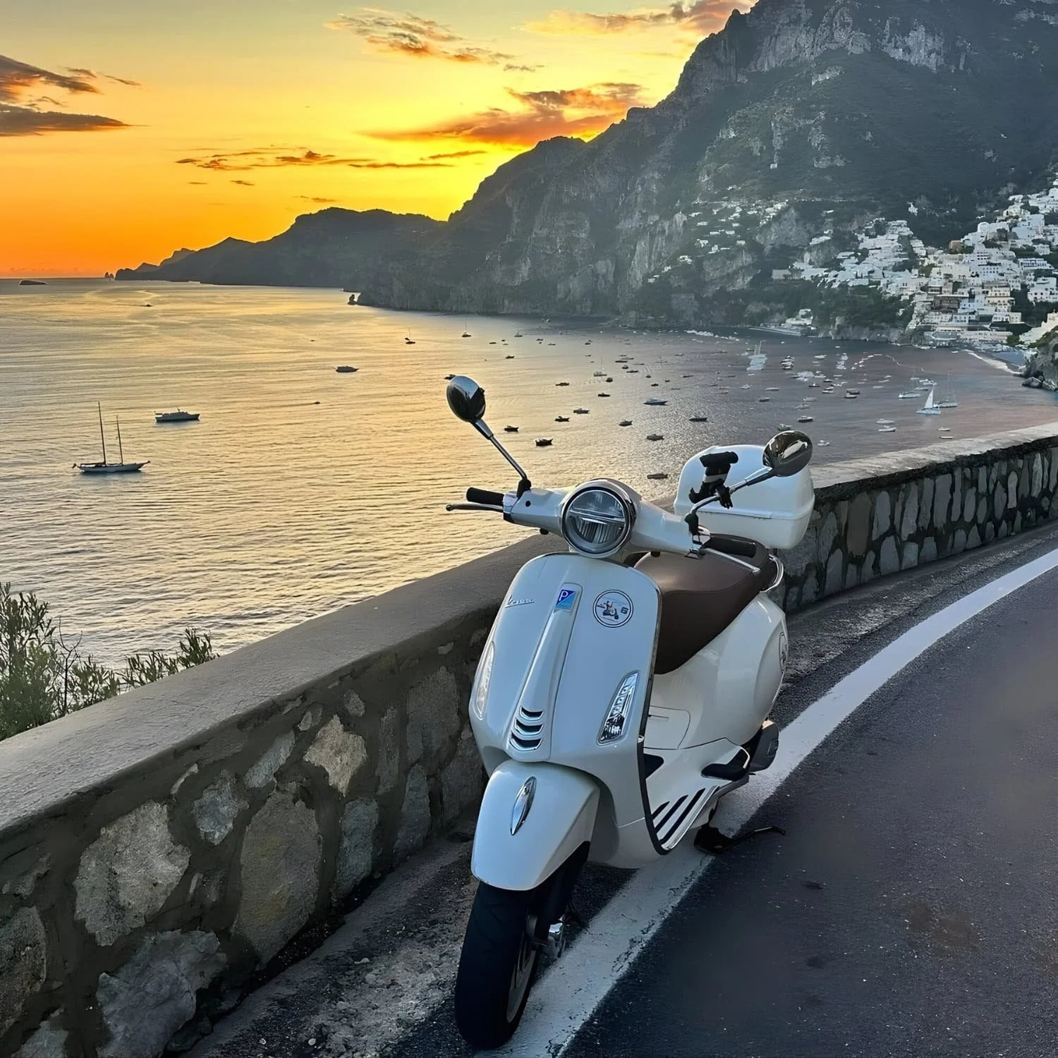 White Vespa parked along the coastal road overlooking Positano at sunset with boats scattered across the bay below. Driving the scenic coastal road is one of the iconic Amalfi Coast things to do.