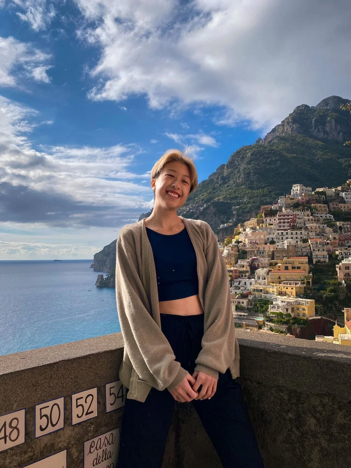 Gi Shieh, travel blogger smiling in front of Positano’s pastel hillside buildings and the blue Mediterranean Sea. Mountains and scattered clouds create a dramatic backdrop along the Amalfi Coast.
