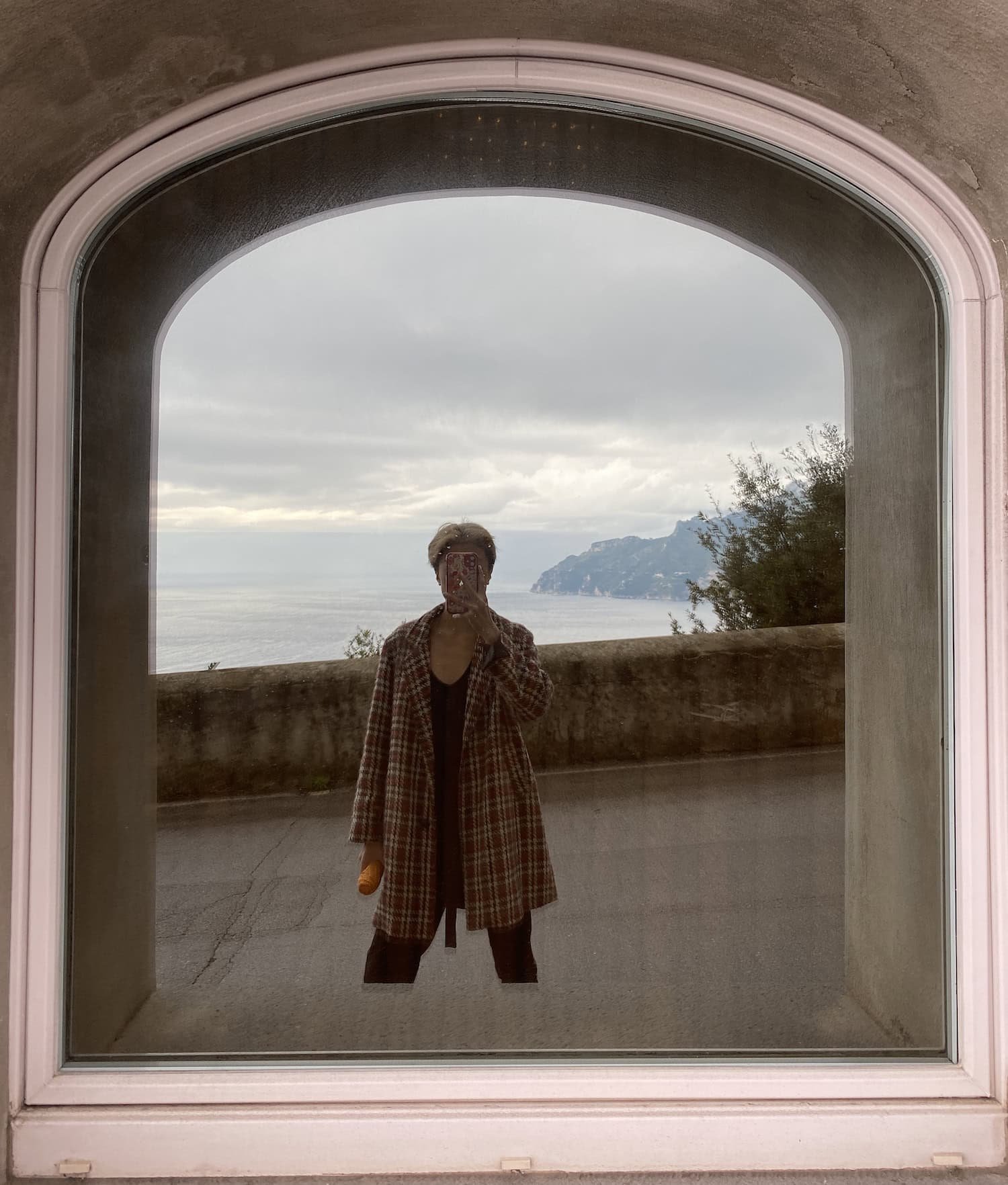 Person taking a mirror selfie through an arched window overlooking the Amalfi Coast with cloudy skies and calm sea in the background. The framing highlights the scenic coastal view often considered when deciding where to stay in Amalfi.
