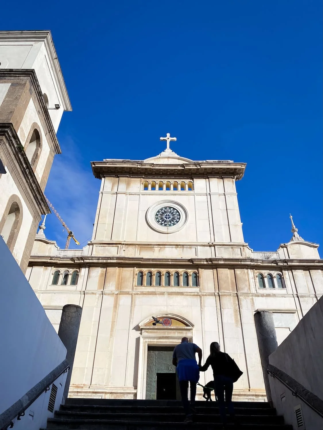 White church facade in Positano photographed against a deep blue sky. Two people walk up the staircase toward the entrance beneath the large cross and circular window.