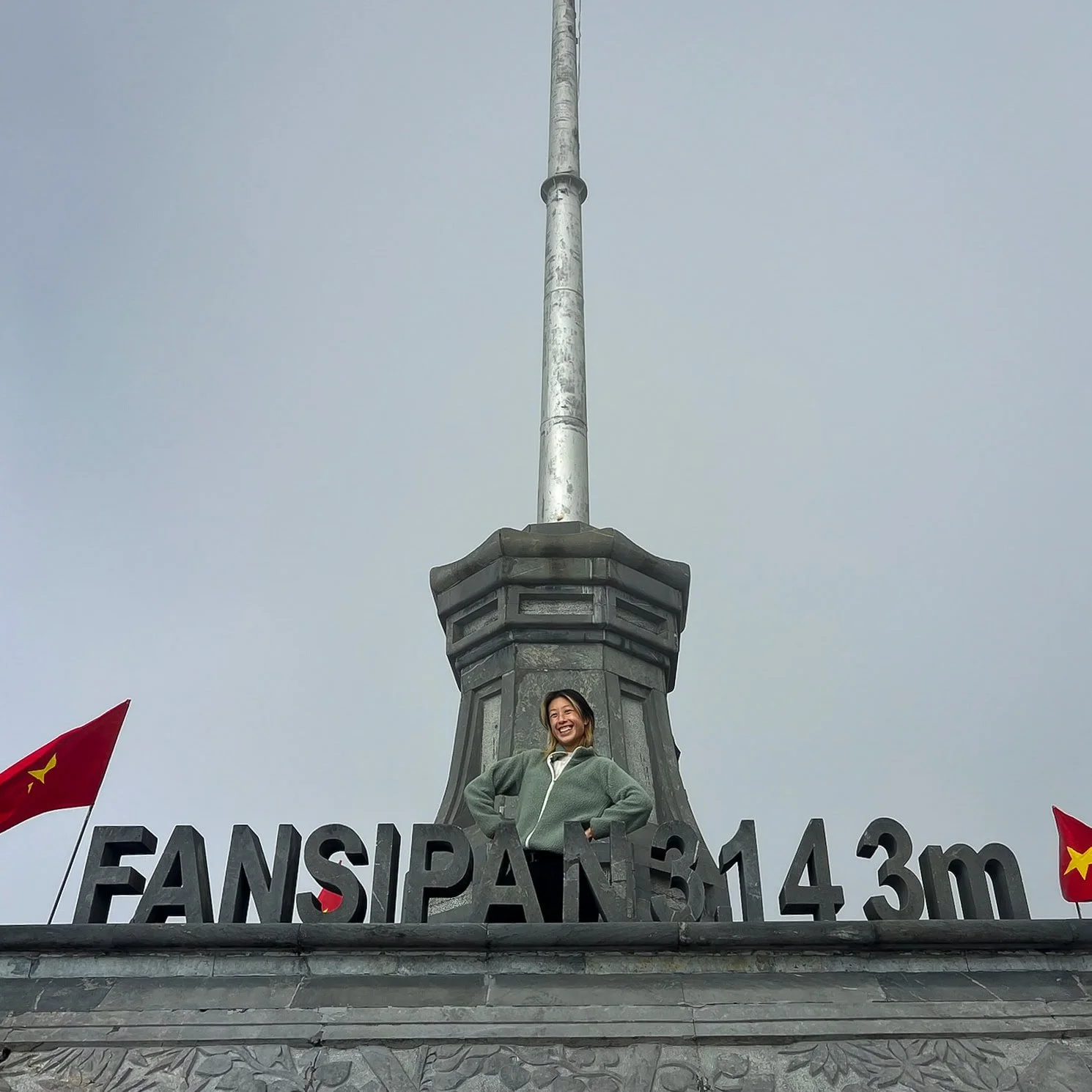 A smiling visitor stands behind the large black sign reading "FANSIPAN 3.143m" at the summit marker of Fansipan, Vietnam’s highest peak, with Vietnamese flags on both sides and a tall silver spire rising above