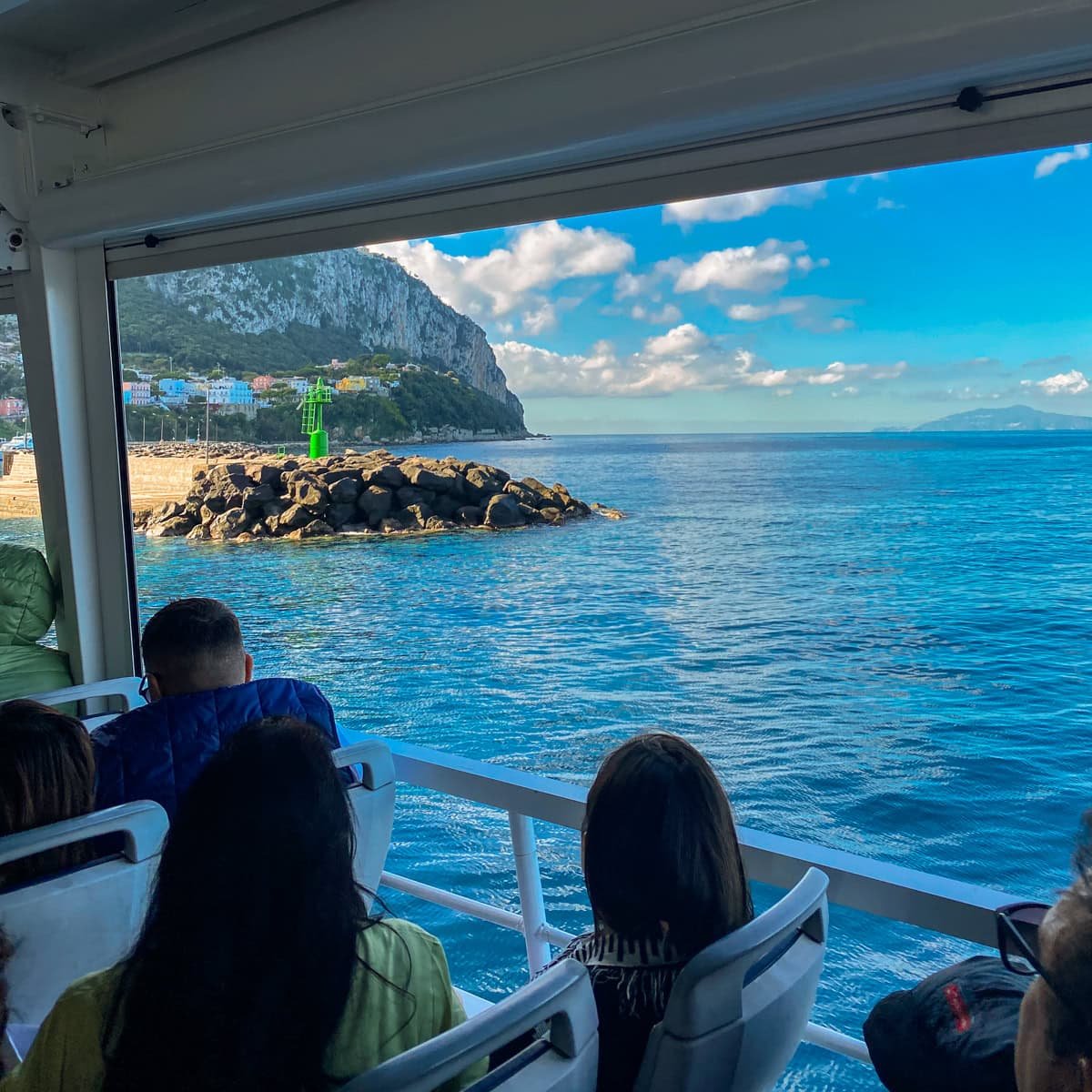 Passengers inside a ferry arriving at Capri, looking at the rocky coastline, a green navigation beacon, and the deep blue sea surrounding the island.