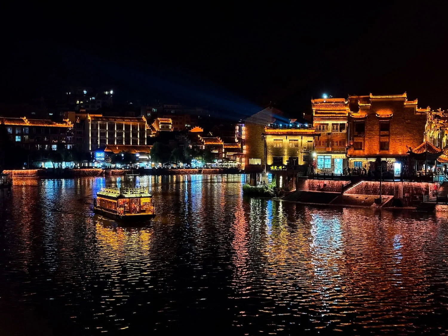 Nighttime view of a river with illuminated traditional buildings and a sightseeing boat. Eastogo includes beautiful evening scenery that showcases China's historic towns after dark.