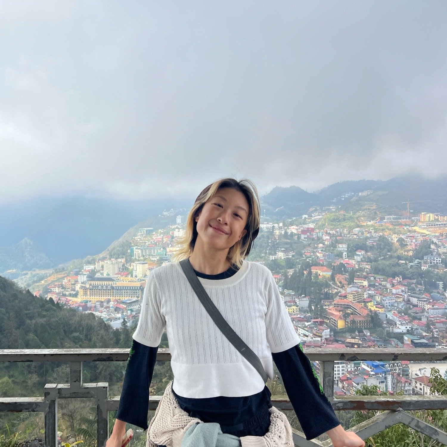 East Asian traveler wearing a white shirt and lots of layers standing in front of Sapa Town, where it’s best to stay in a homestay.
